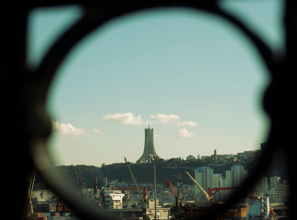 View of Algiers through a circular frame, with the Maqam Echahid memorial visible on the hillside above the port